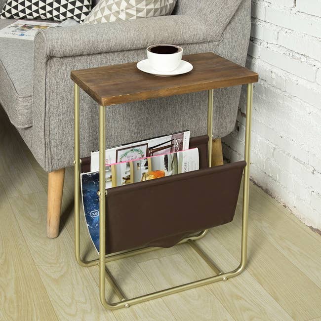 Side table with a wooden top, metal legs, and a magazine rack underneath, holding magazines and a cup of coffee on top, next to a sofa