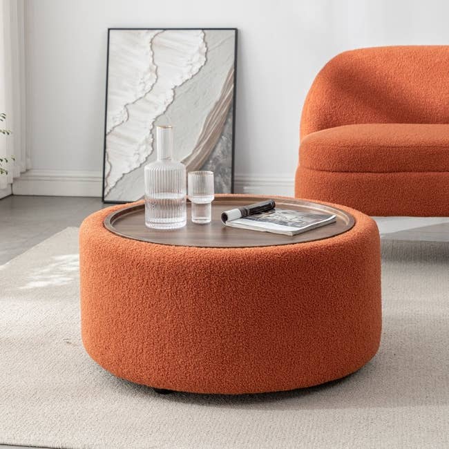 Round orange ottoman with a wooden tray top, set with a glass carafe and tumbler, in a modern living room. Abstract art leans against the wall