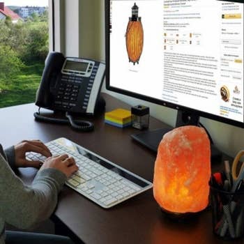 Person shopping online at a desk with a lit Himalayan salt lamp, telephone, keyboard, and monitor displaying a product page