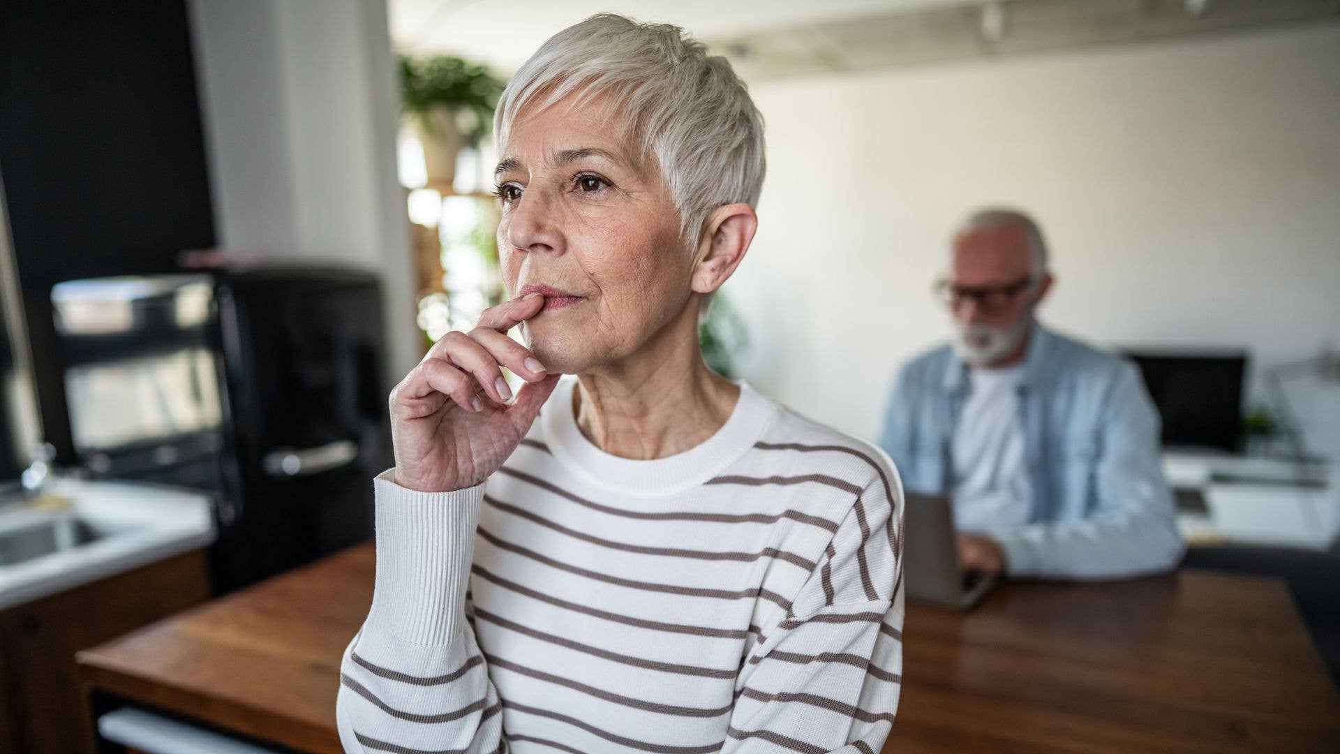 Senior woman looking pensive and thoughtful, with her hand on her chin, while a man works on a laptop in the background, possibly in their home or apartment