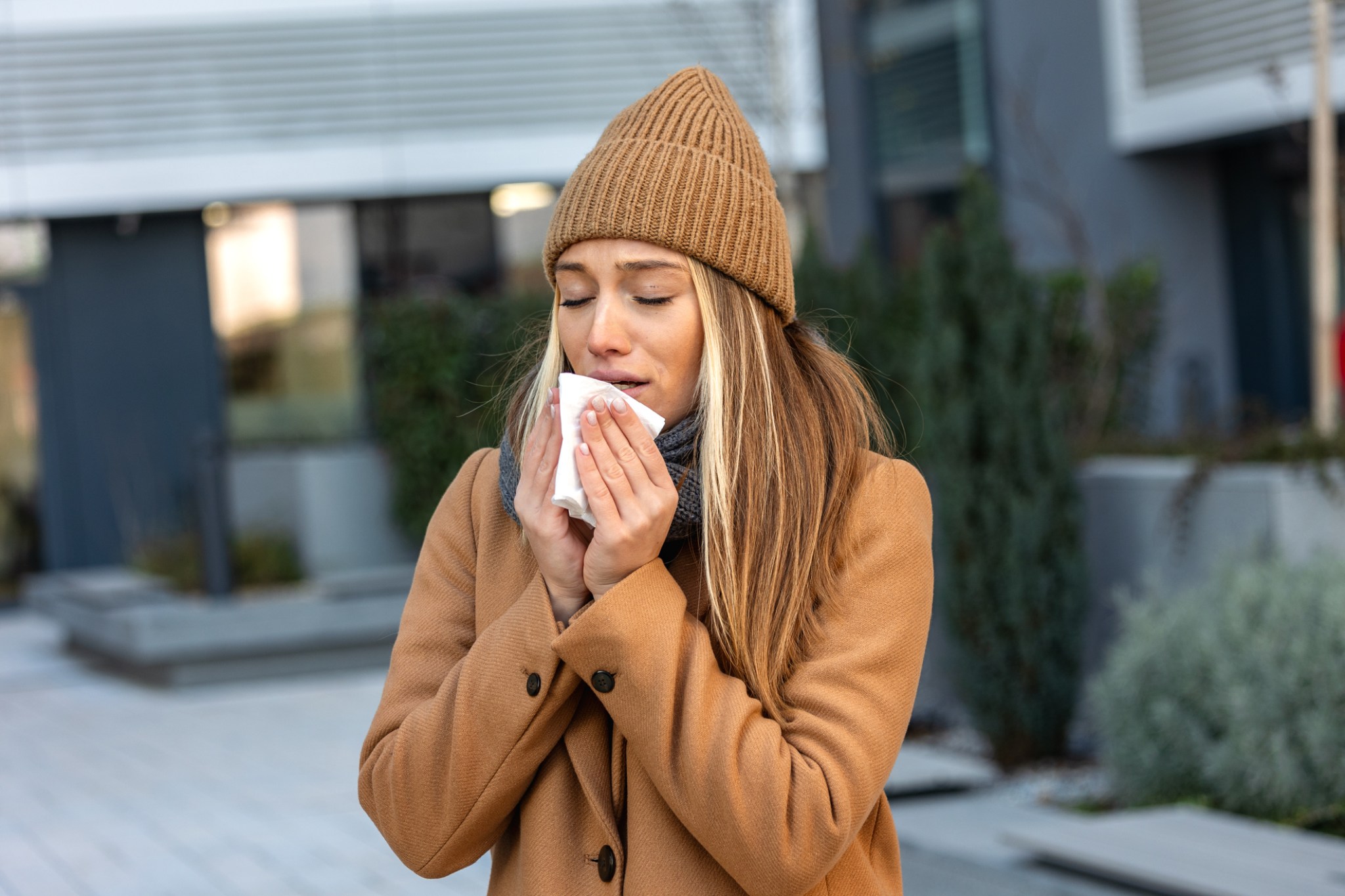 Female Blowing Nose with Tissue During Winter Cold. Woman wearing winter clothes sneezing or blowing her nose with a tissue in an urban setting. Represents flu symptoms, allergies, and health problems