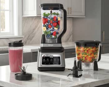 Blender set on a kitchen counter with fruits inside, smoothie in a to-go cup, chopper with diced vegetables, and blender attachments nearby
