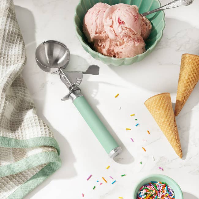 Ice cream scoop with a green handle next to a bowl of strawberry ice cream, empty cones, colorful sprinkles, and a textured towel