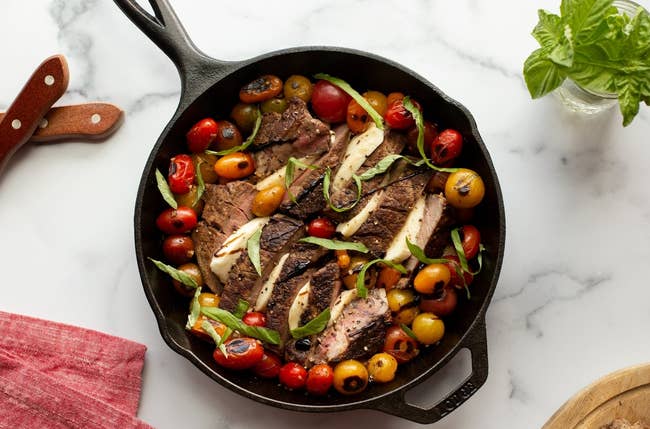 Skillet with grilled steak, cherry tomatoes, and herbs on a marble surface. Knife, napkin, and basil nearby