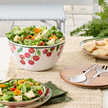 A set of fruit-patterned salad bowls and serving utensils, with fresh salad and bread arranged on a woven placemat, ideal for outdoor dining