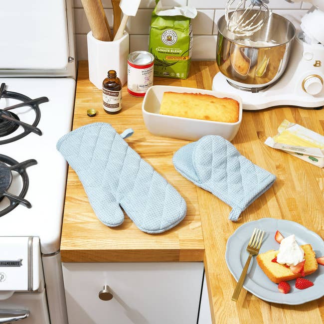 Two quilted oven mitts on a kitchen countertop beside a loaf of cake, mixer, and ingredients. A plated slice is garnished with strawberries