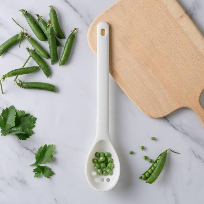 White spoon with peas, parsley, and a cutting board on a marble surface. Ideal for cooking or kitchen preparation articles