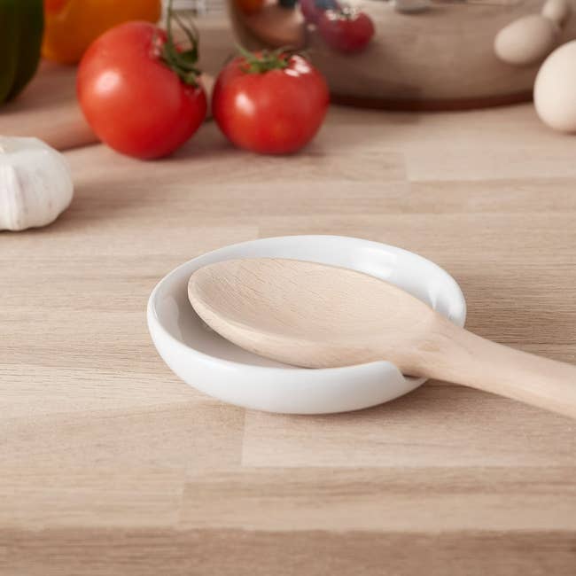 Wooden spoon rests on a white ceramic dish, surrounded by fresh vegetables and kitchen items, suggesting a cooking or kitchen shopping theme
