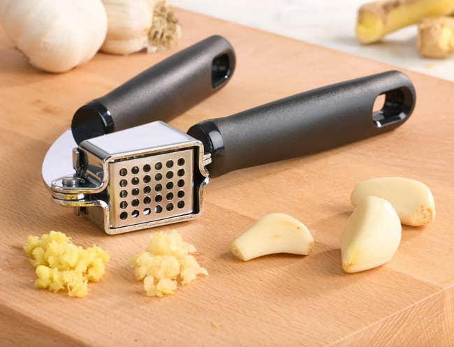 Garlic press on wooden board with crushed garlic and garlic cloves nearby; kitchen tool for easy garlic preparation