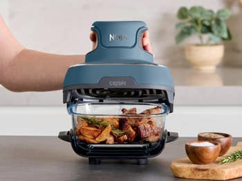 A hand operates a Ninja air fryer with roast beef and potatoes inside, placed on a kitchen counter