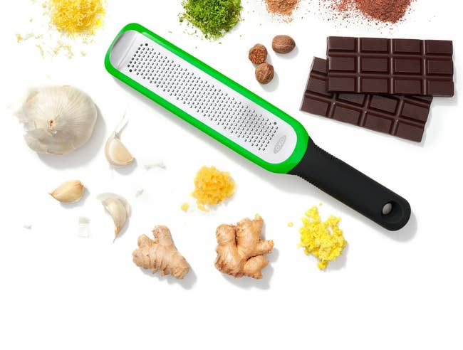 Grater with handle surrounded by spices, chocolate, garlic, and ginger, showcasing kitchen essentials for enhancing flavors in cooking
