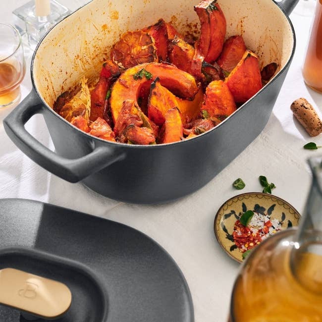 Cast iron Dutch oven filled with roasted squash and herbs, accompanied by a bottle of olive oil and small plate with spices on a white tablecloth
