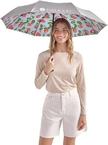 Smiling person holds an umbrella with a strawberry pattern, wearing a casual long-sleeve top and shorts