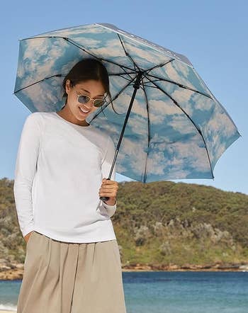 Person on a beach holding an umbrella with a cloud pattern, wearing sunglasses, a long-sleeve shirt, and wide-leg pants