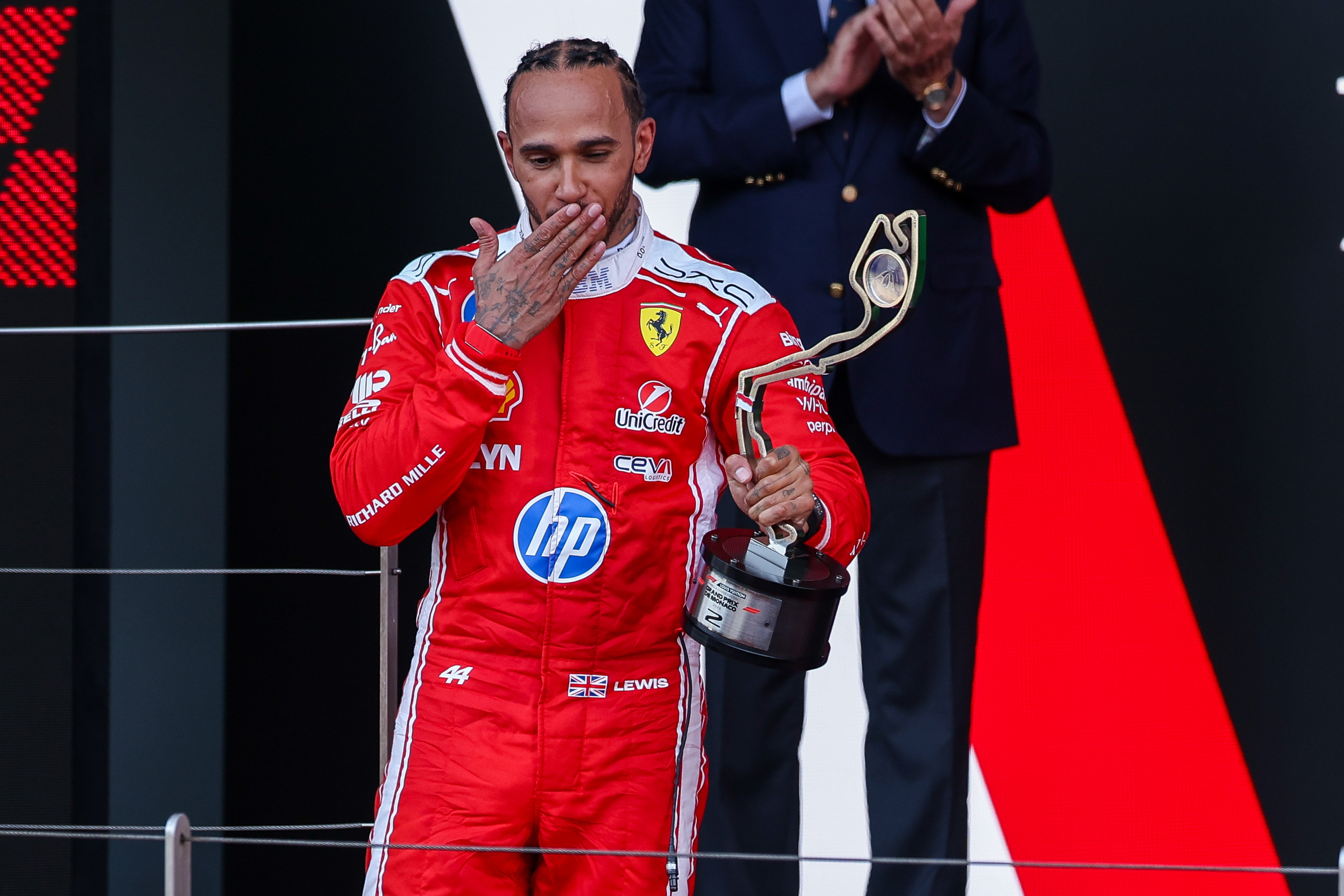 Lewis Hamilton celebrates on the podium, holding a trophy shaped like a racing track, and blowing a kiss to the crowd.