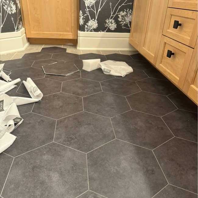Hexagonal floor tiles arranged in a pattern with additional tiles and wrapping materials scattered on the floor. Wood cabinetry in the background