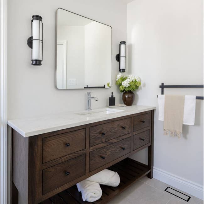 Modern bathroom vanity with a wooden cabinet, white countertop, rectangular mirror, and contemporary lighting. Towels and flowers are neatly placed