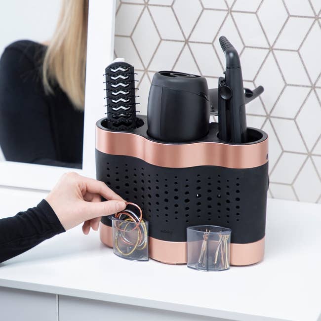 Hair styling tool organizer with compartments for a hair dryer, straightener, brush, and hair accessories; person placing hair ties inside