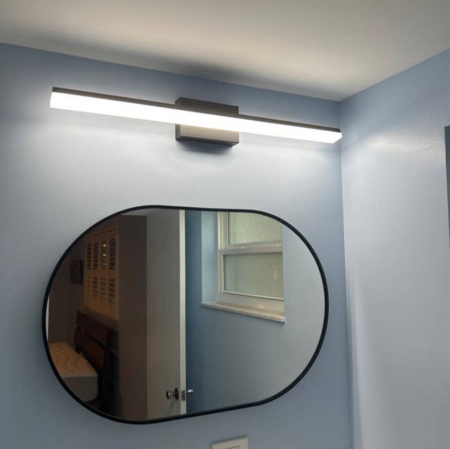 Oval mirror under a modern horizontal light fixture in a bathroom with a window reflection
