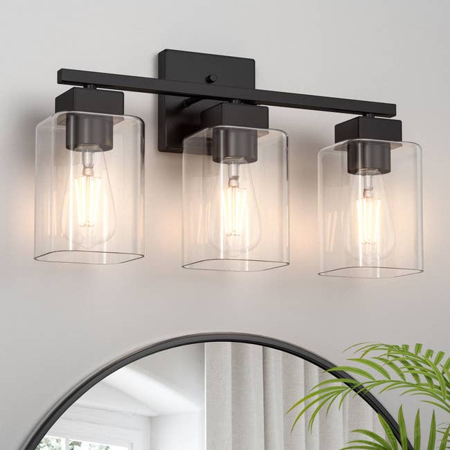 Three clear glass cylinder wall sconces with visible filament bulbs, mounted on a rectangular black base above a round mirror and plant. Ideal for modern decor