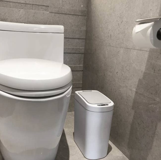 Modern bathroom showing a white toilet and a matching small waste bin with a lid next to it. There is a toilet paper holder affixed to the wall