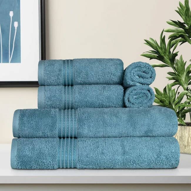 Stack of blue towels of varying sizes displayed on a white surface with a framed picture and a plant in the background