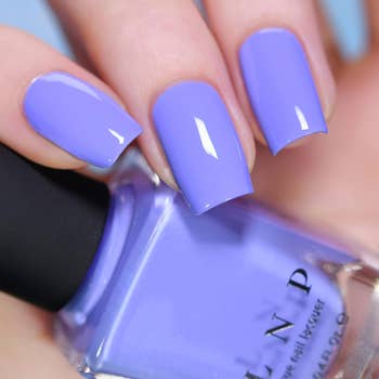 Close-up of manicured nails painted with glossy lavender nail polish, bottle held in hand. Featured in a shopping article