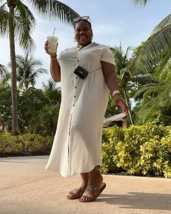 Person outdoors in a button-up dress holds a drink, smiling under palm trees. They wear sunglasses and sandals, with a small crossbody bag