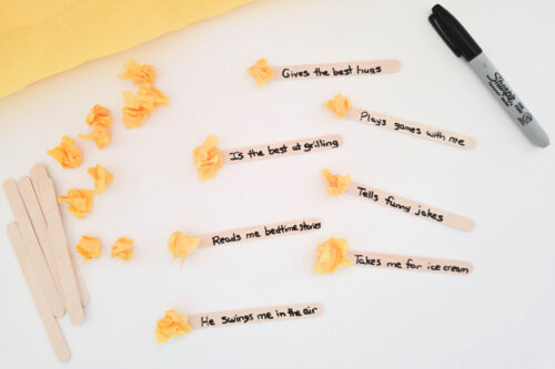 Several craft sticks with handwritten messages such as "Gives the best hugs" and "Takes me for ice cream" are arranged on a table with a marker and crumpled yellow paper.