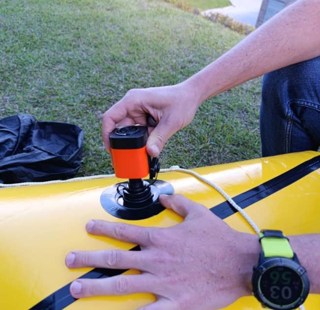Hand inflating a yellow inflatable item outdoors with an air pump. A smartwatch is visible on the wrist