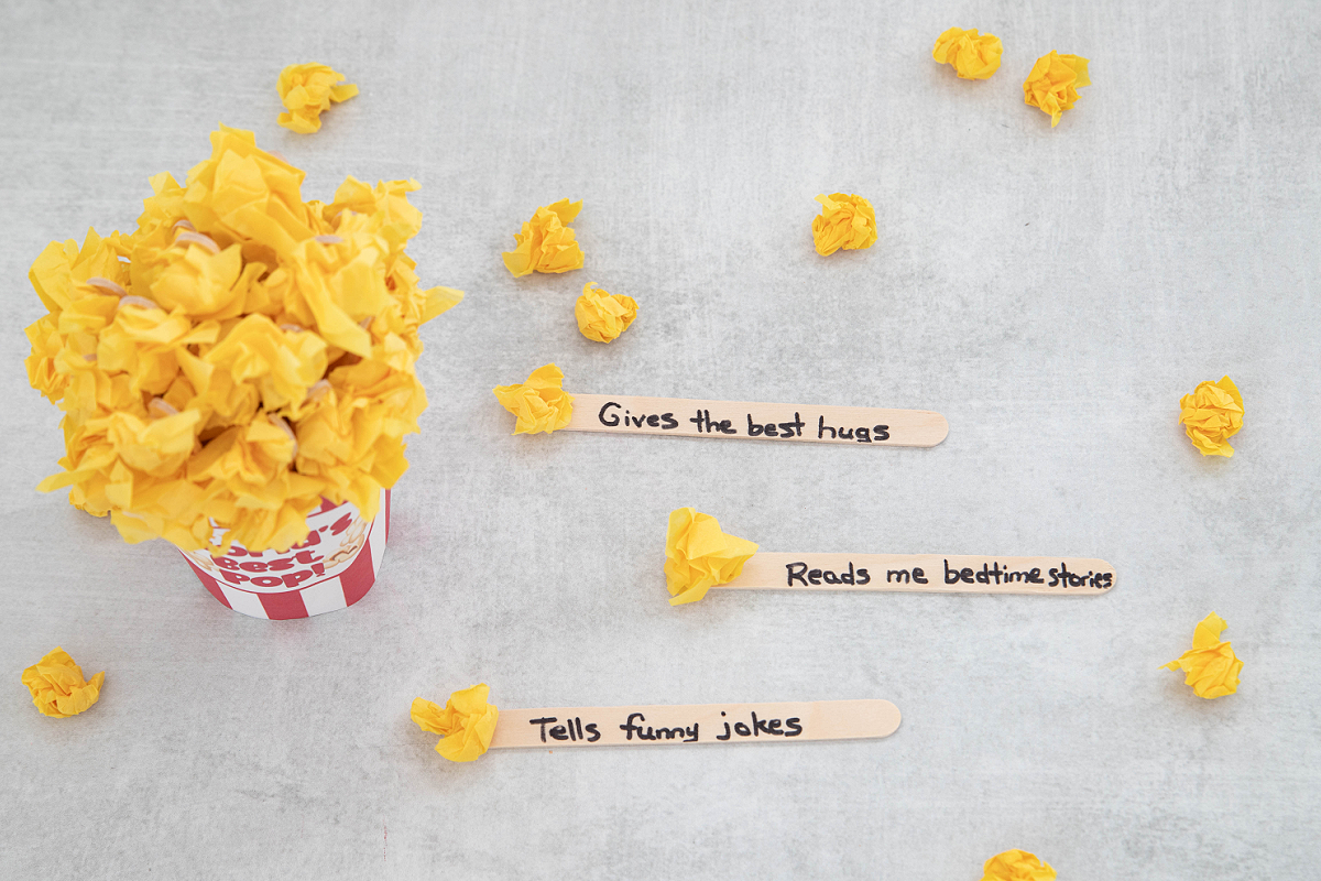 A popcorn container filled with yellow paper sits on a gray surface, with three craft sticks labeled: “Gives the best hugs,” “Reads me bedtime stories,” and “Tells funny jokes.”.