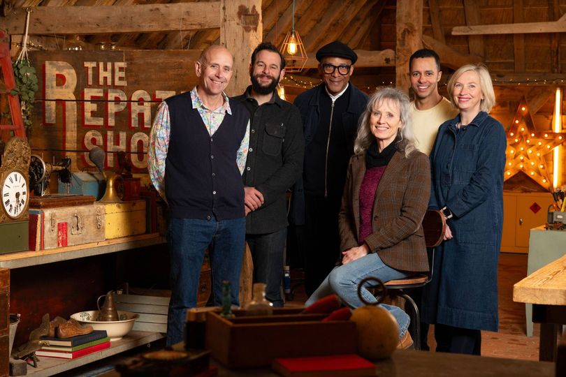 A group of individuals dressed in winter clothing, including coats and vests, are gathered in a workshop setting. They stand beside a wooden structure with various shelves and items, creating an environment that suggests craftsmanship or DIY activities. The setting is warmly lit, and the individuals appear to be posing for a photograph.