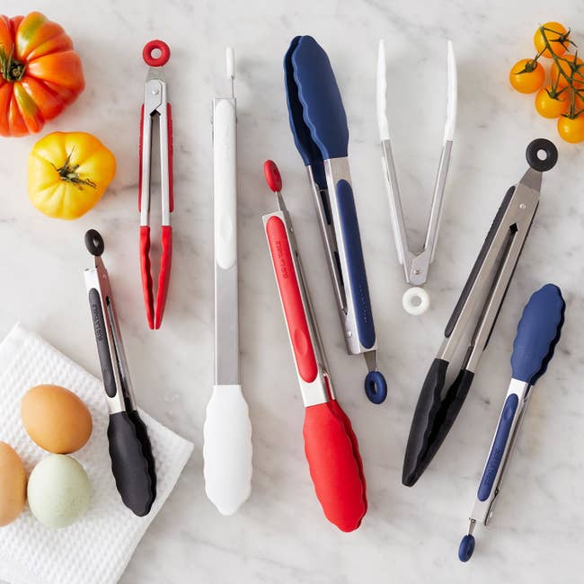 Assorted kitchen tongs with silicone grips are displayed on a marble surface with tomatoes and eggs nearby
