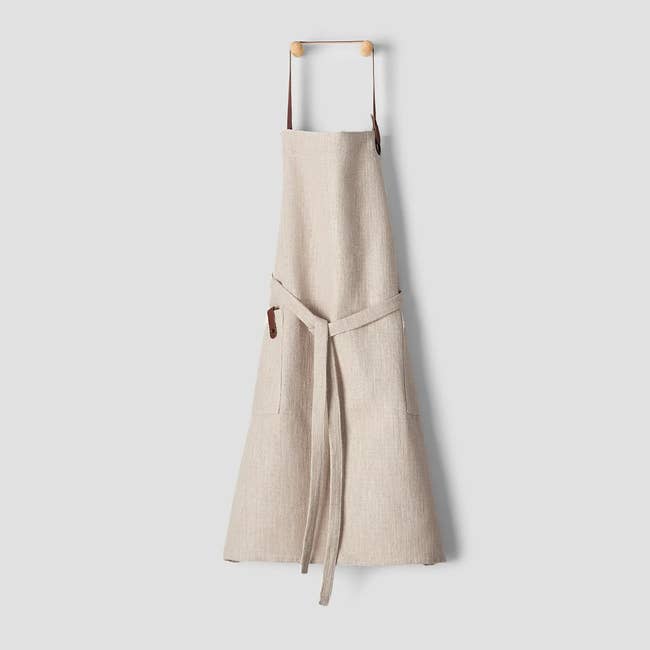 Beige linen apron hanging on a hook with a front pocket and adjustable straps. Perfect for cooking or baking enthusiasts