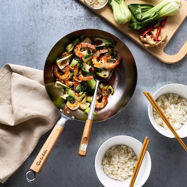 A stir-fry with shrimp and vegetables in a wok, surrounded by bowls of rice and fresh ingredients on a cutting board, designed for a shopping feature