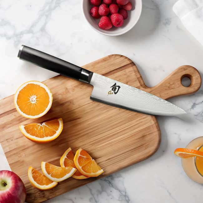 A chef's knife on a wooden cutting board with sliced oranges, an apple, strawberries, and a glass of orange juice nearby
