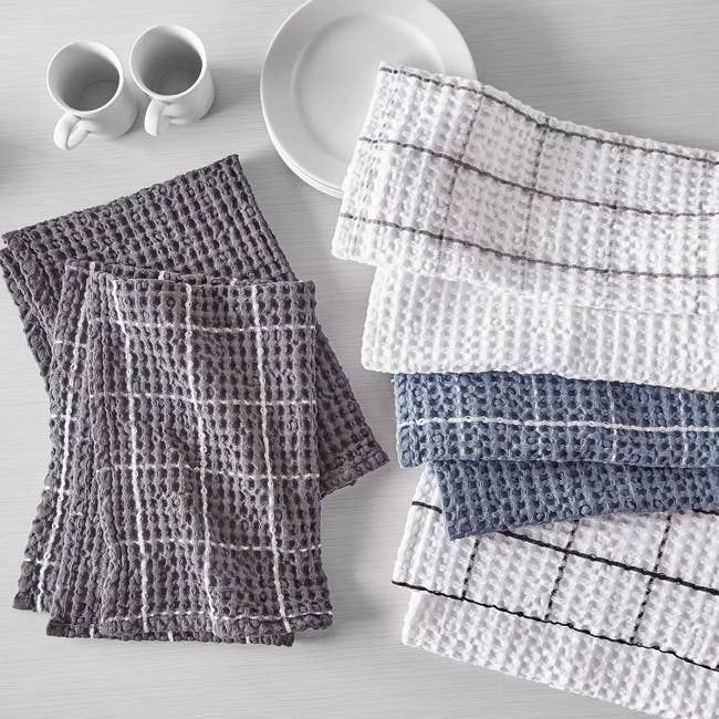A set of waffle-patterned kitchen towels in white, gray, and blue, displayed next to stackable white dishware and mugs on a countertop