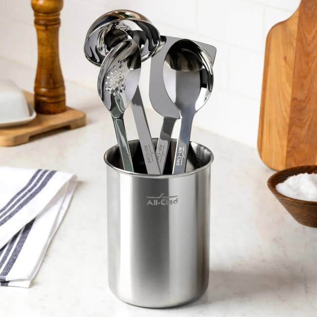 Set of stainless steel kitchen utensils in a metal holder on a countertop, with pepper mill and salt dish nearby
