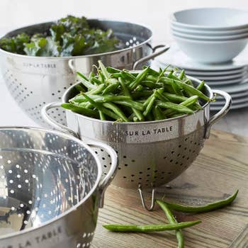 Metal colanders with green beans and greens. Stacked white bowls in the background. Image suggests kitchenware for sale