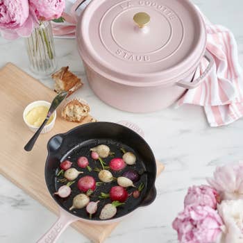 A Staub cast iron skillet with radishes next to a matching pink cocotte, surrounded by bread and flowers on a kitchen counter