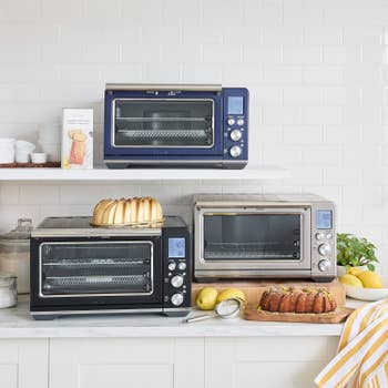 Three countertop ovens are displayed on white shelves. Freshly baked cakes, lemons, and kitchen items surround them, suggesting a baking theme