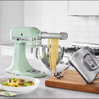 Countertop with a stand mixer rolling pasta, surrounded by a hand mixer and a plate of prepared pasta. Kitchen setting