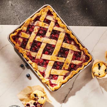 Rectangular berry pie with lattice crust, surrounded by three muffins on a marble surface