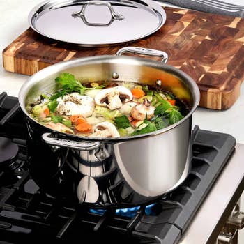 Soup with vegetables and mushrooms simmers in a stainless steel pot on a stove next to a wooden cutting board