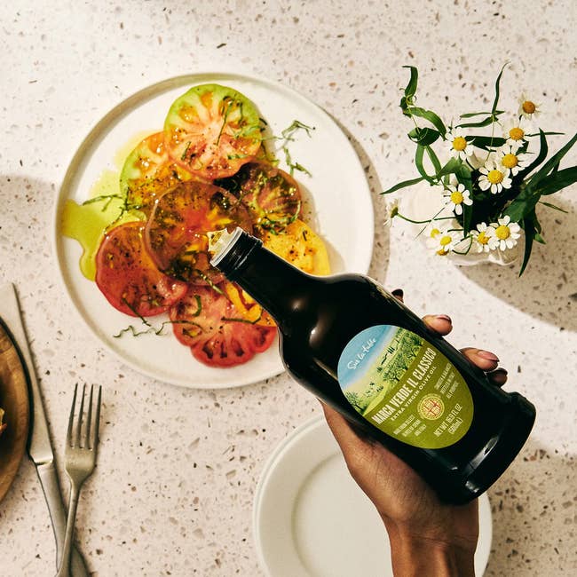 Hand holding a bottle of walnut oil over a plate of sliced tomatoes with herbs, placed on a table with a fork, knife, and flowers