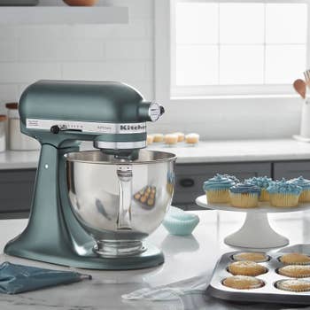 A stand mixer on a kitchen counter near freshly baked cupcakes, some with blue frosting. There's a cupcake pan beside it