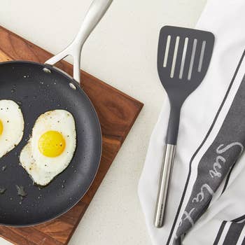 Frying pan with two sunny-side-up eggs on a wooden cutting board, next to a black spatula and a striped kitchen towel