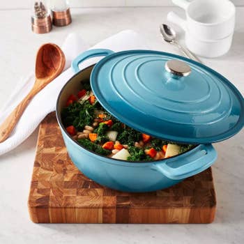 A blue cast iron pot with a partially open lid shows a hearty vegetable and bean stew, placed on a wooden cutting board in a kitchen setting