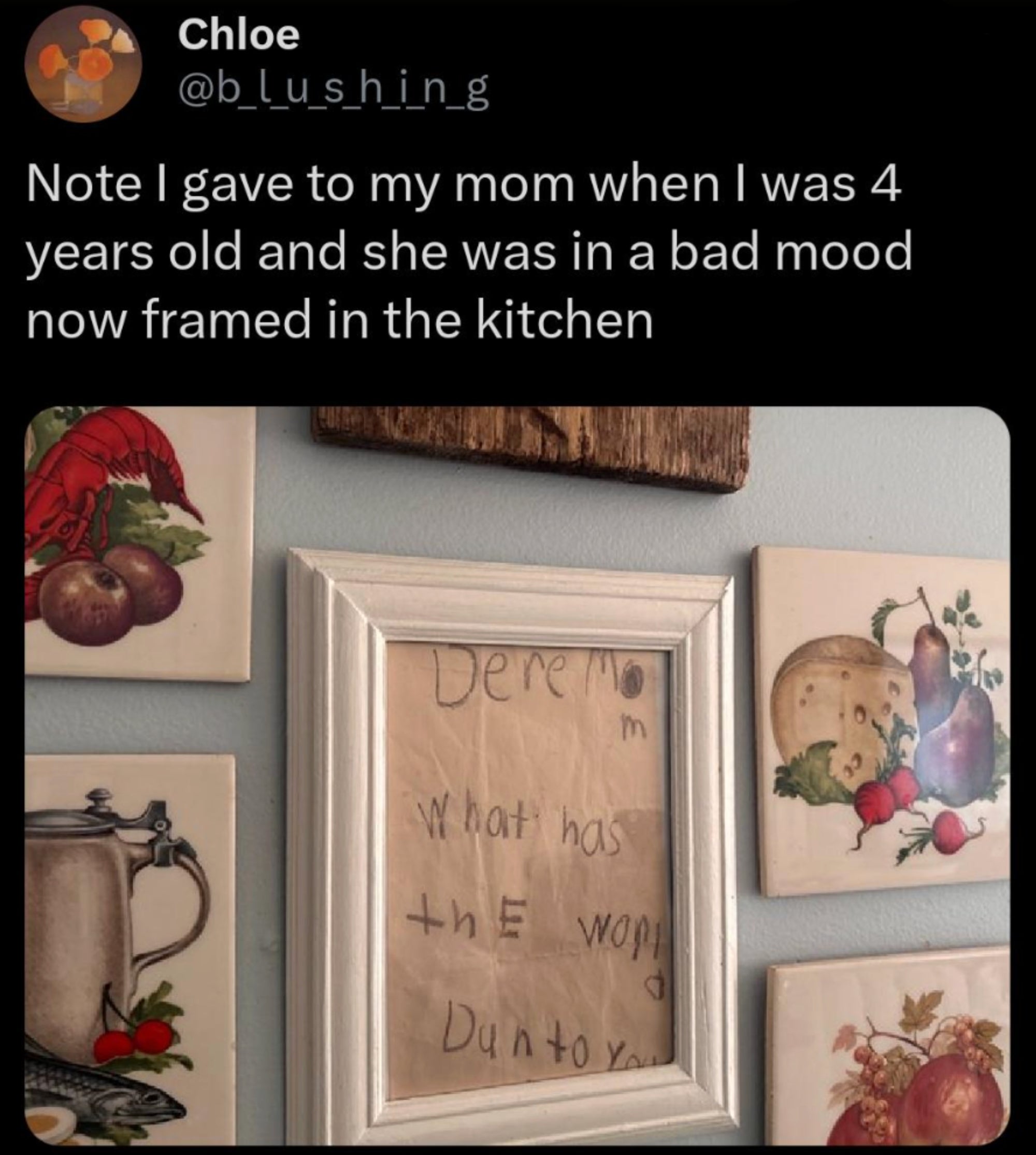 Framed note from a child to their mom, humorously displaying early handwriting, hangs in a kitchen, surrounded by art pieces of food