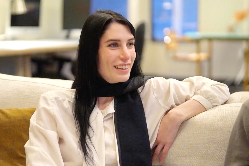 Georgia McCann wearing a black scarf and white shirt while sitting on a couch.